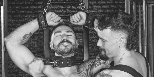 An erotic image of a BDSM scenario, featuring a booted foot on a mans chest. The mans wearing a leather cocksucker hood.